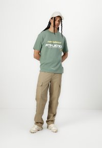 Zaļš kokvilnas T-krekls ar baltu un dzeltenu "new balance ATHLETICS" zīmolu, kombinēts ar brūnām kravas biksēm un krēmkrāsas kedām.