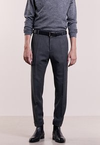 Hombre vestido con pantalones de traje grises, cinturón negro, camisa de manga larga gris y botas de vestir de cuero negras, de pie sobre un suelo claro.