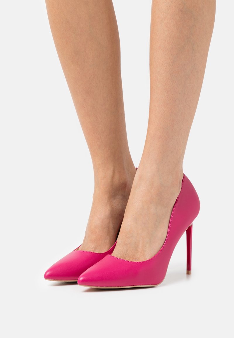Jambes portant des escarpins à bout pointu couleur rose vif avec talons aiguilles sur un fond blanc uni.