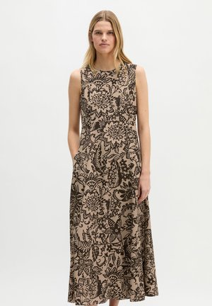 Ärmelloses beige Maxikleid mit schwarzem, aufwändigem Blumenmuster, getragen von einer stehenden Frau mit langen blonden Haaren vor einfarbigem Hintergrund.