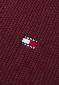 Burgundia ribade kangas, millel on kudumine plaastriga "TOMMY JEANS" valge ja tumesinise värviga, tähistades punase ja valge horisontaalsete triipudega.
