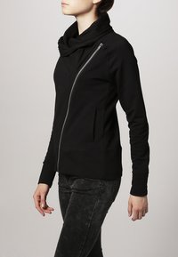 Svart huvtröja med dragsko, asymmetrisk dragkedja och sidofickor. Tillverkad av mjukt tyg med en figurnära passform.