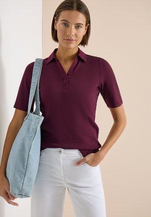 Kvinde iført en vinrød poloshirt med korte ærmer og hvide bukser, bærende en stor lyseblå tote-taske over skulderen.