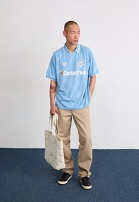 Light blue short-sleeve jersey with white accents, logo on the chest, paired with beige pants and a cream quilted tote bag. Black shoes.