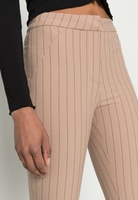 Calças bejes com riscas verticais, feitas de um tecido suave e com uma forma afunilada. Apresentam uma cintura plana e detalhes de bolsos laterais, mostradas com uma mão.