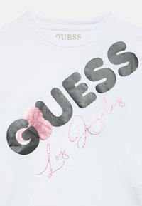 Άσπρο βαμβακερό T-shirt με μεγάλα μαύρα γράμματα "GUESS", με ροζ γκλίτερ καρδιά και ροζ κείμενο με καλλιγραφική γραφή "Los Angeles" από κάτω.