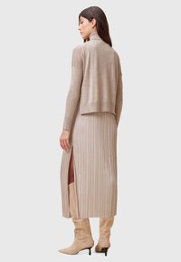 Maglione beige corto sopra un lungo abito a pieghe beige con spacchi laterali, abbinato a stivali beige sopra il ginocchio. Texture liscia in tutto il look.