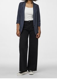 Persona che indossa un blazer blu navy, una maglietta bianca, jeans neri a gamba larga e sneakers platform nere, in piedi contro uno sfondo chiaro e uniforme.