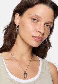 Sølv kædedyse med en iøjnefaldende sikkerhedsnål vedhæng, båret over en lys beige tank top med et hvidt inderlag.