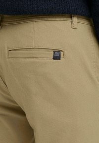 Pantalons beiges avec une poche arrière portant une petite étiquette noire, confectionnés dans un tissu texturé, avec des détails de couture visibles.