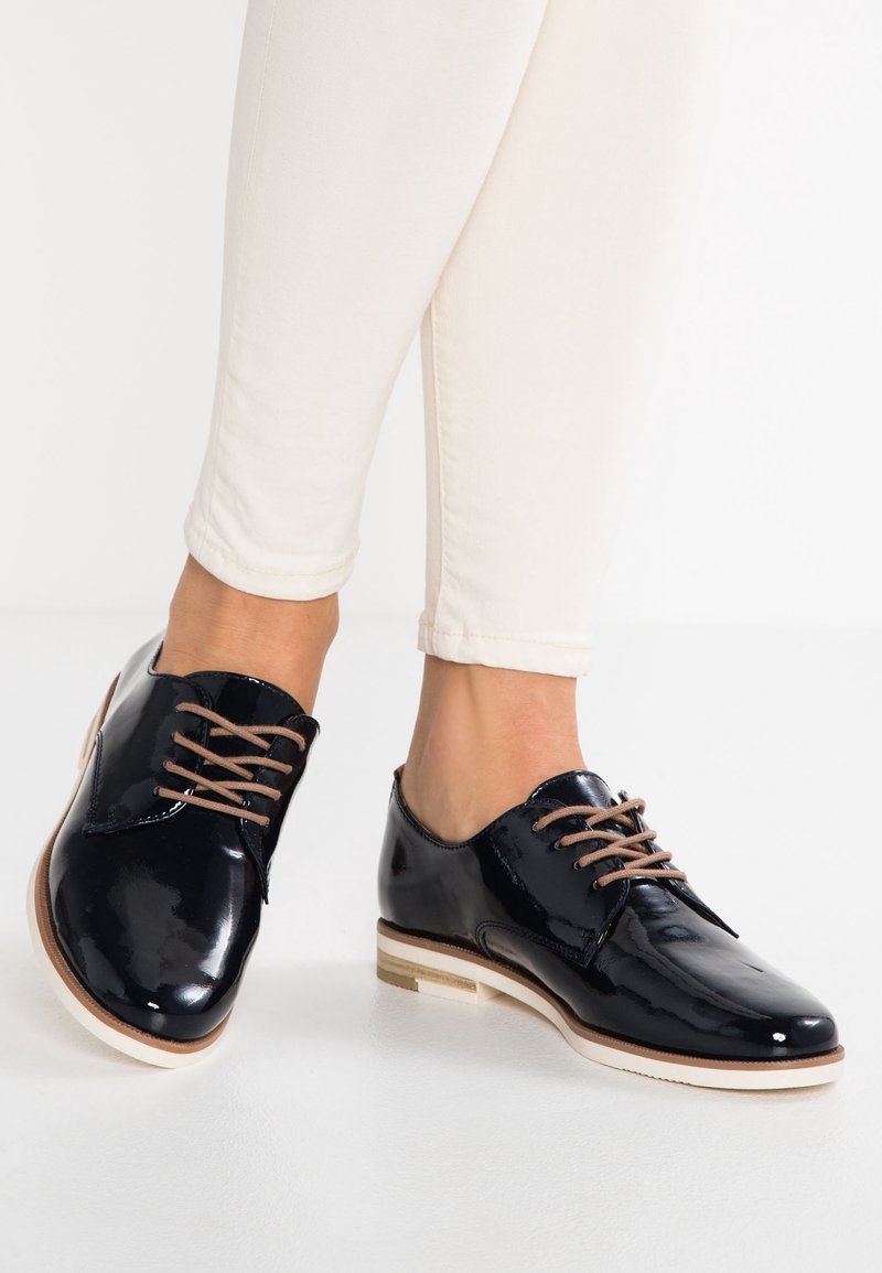 Chaussures Oxford noires brillantes à lacets avec semelles beiges portées avec un pantalon blanc ajusté, présentées sur un fond blanc uni.