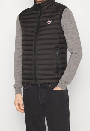 Homme portant un gilet matelassé noir sans manches sur une chemise grise à manches longues et un jean noir, une main dans la poche.