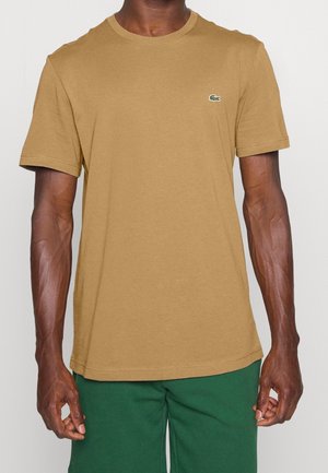 Homme portant un t-shirt marron uni à manches courtes avec un petit logo crocodile vert sur la poitrine, associé à un pantalon vert.