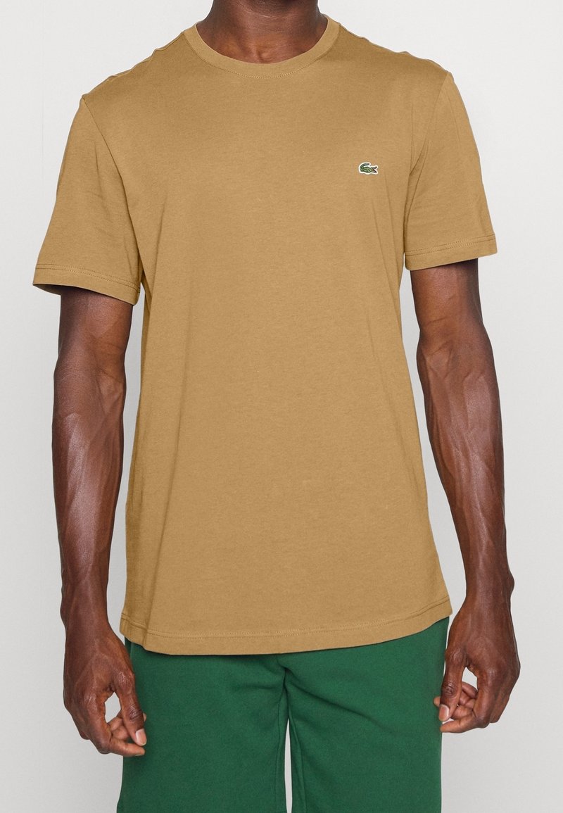Homme portant un t-shirt marron uni à manches courtes avec un petit logo crocodile vert sur la poitrine, associé à un pantalon vert.