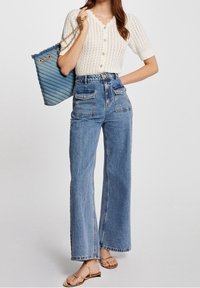 Cárdigan blanco de punto con escote en forma de onda, jeans de pierna ancha de talle alto en azul, bolso de cadena dorado y sandalias planas doradas.