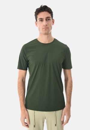 Uomo che indossa una maglietta verde a maniche corte e pantaloni beige, in piedi contro uno sfondo chiaro e uniforme, rivolto in avanti con un'espressione neutra.