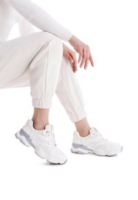 Zapatillas deportivas blancas con tejido texturizado, detalles de malla y suelas de goma gris. Combinadas con pantalones de chándal de color claro, presentan un aspecto deportivo.