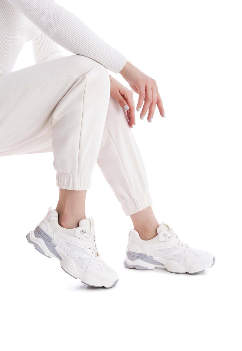 Zapatillas deportivas blancas con tejido texturizado, detalles de malla y suelas de goma gris. Combinadas con pantalones de chándal de color claro, presentan un aspecto deportivo.