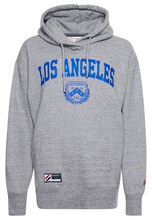 Gros plan d'une personne portant un sweat à capuche gris avec le texte bleu « LOS ANGELES » et un emblème bleu avec le texte « SUPERDRY 1954 CAL ».
