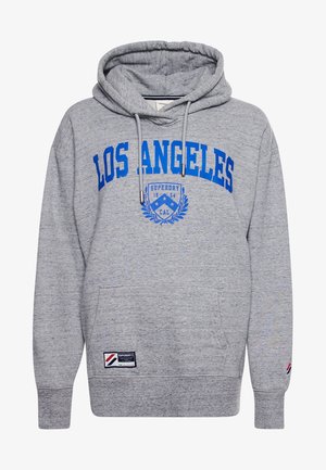 Close-up van een persoon die een grijze hoodie draagt met blauwe tekst "LOS ANGELES" en een blauwe embleem met de tekst "SUPERDRY 1954 CAL".