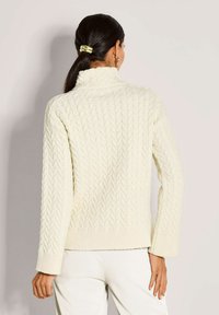 Vrouw met donker haar in een staart draagt een crèmekleurige kabelgebreide coltrui en witte broek, gezien vanuit de rug tegen een eenvoudige achtergrond.
