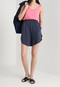 Różowy top na ramiączkach w połączeniu z ciemnoszarymi szortami, posiadającymi elastyczny pasek i luźny krój. Czarne klapki dopełniają stylizację.