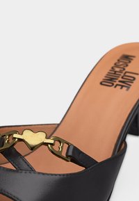 Sandale noire à talon haut avec détail de chaîne dorée en forme de cœur et semelle intérieure beige marquée "LOVE MOSCHINO" sur fond uni.