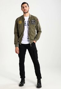 Olijfgroene bomberjack met patches, ritssluiting en geribbelde manchetten, gedragen over een wit t-shirt en zwarte cargobroek met zakken.