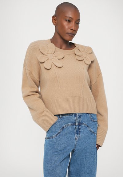 Beige stickad tröja med överdimensionerade blommorapplikationer på bröstet, ribbad mudd och kortad nederkant, ihop med lösa blåjeans.