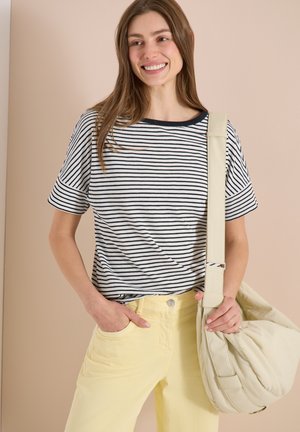 Femme souriante portant une chemise rayée noir et blanc et un pantalon jaune, tenant un sac à bandoulière crème sur fond beige.