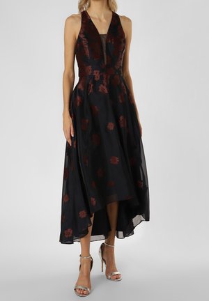 Frau trägt ärmelloses schwarzes Kleid mit roten Blumenmustern, tiefem V-Ausschnitt, asymmetrischem Saum und silbernen High-Heel-Sandalen mit Knöchelriemen.