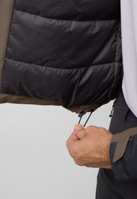 Geïsoleerde jas in bruin en zwart, met een gedeeltelijk open ritssluiting aan de binnenkant, een puffy textuur en verstelbare trekkoorden aan de onderkant.