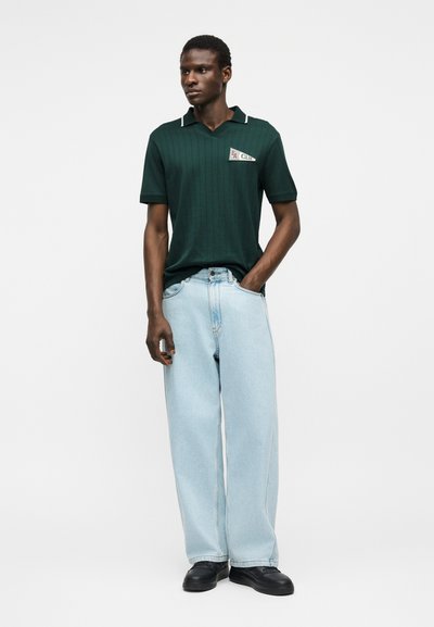 Model męski ubrany w ciemnozieloną koszulę z krótkim rękawem z naszywką "EA Club", szerokie jasnoniebieskie dżinsy oraz czarne buty, stojący na białym tle.