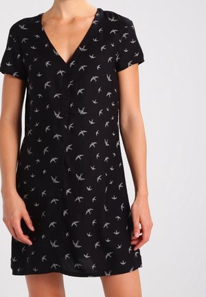 Black short-sleeve dress with V-neck and all-over white bird print, worn by a person standing with hands relaxed at sides.