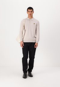 Polo grigio chiaro a maniche lunghe con colletto e logo giallo sul lato sinistro del petto; abbinato a pantaloni neri e scarpe nere; tiene una borsa a spalla beige.