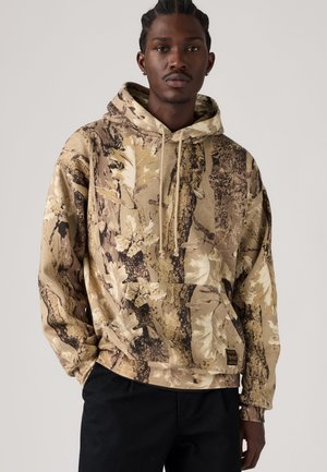 Jeune homme portant un sweat à capuche beige et marron à motif camouflage avec un pantalon noir sur fond uni.