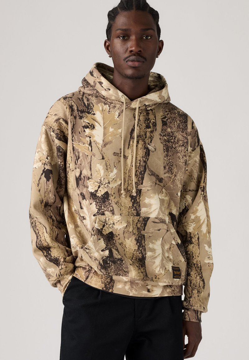 Jeune homme portant un sweat à capuche beige et marron à motif camouflage avec un pantalon noir sur fond uni.