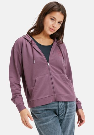 Purple zip-up hoodie made of soft fabric, featuring a hood, front pockets, and adjustable drawstrings. Model wears it with a black top.
