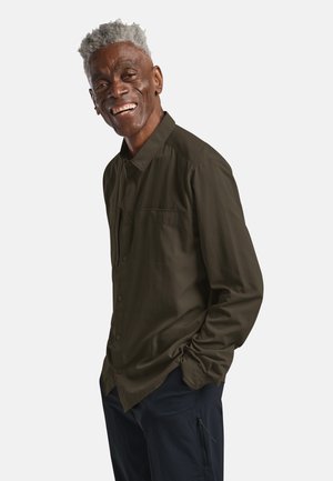 Lächelnder älterer Mann mit grauen Haaren, der ein dunkelolivfarbenes Hemd mit Knöpfen und marineblaue Hosen trägt, mit den Händen in den Hosentaschen stehend vor einem weißen Hintergrund.