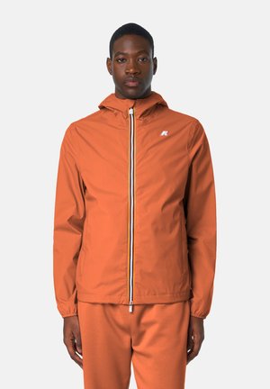 Giovane uomo con giacca arancione con cappuccio e zip e pantaloni abbinati, in piedi davanti a uno sfondo chiaro e uniforme.