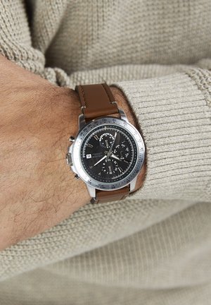 Montre à bracelet en cuir marron avec cadran noir, lunette argentée métallisée et fonctions chronographe. Pull texturé en arrière-plan.