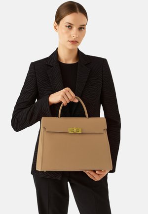 Helle Lederhandtasche mit strukturierter Form, oberen Griff, goldenen Beschlägen und flacher Frontklappe. Das Modell trägt ein schwarze, strukturierten Blazer.