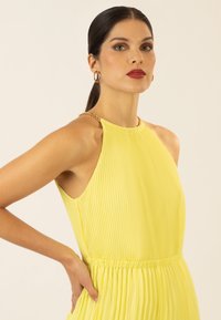 Robe plissée jaune avec un col halter et une taille froncée. Présente un détail de chaîne en or au niveau de l'encolure et une texture de tissu lisse.