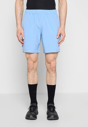 Homme debout portant un short de sport bleu clair, des chaussettes noires et des baskets noires sur un fond gris uni.