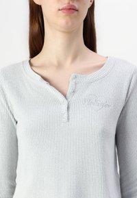 Lichtgrijze geribbelde top met lange mouwen, voorzien van een henley-nek met drie knopen en een subtiele geborduurde hartdetail. Stof lijkt zacht en gestructureerd.