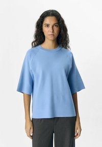 Tee-shirt en tricot bleu à manches raglan courtes, col rond, texture lisse et coupe décontractée, associé à un pantalon gris.