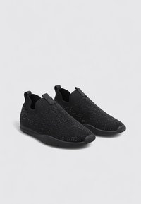 Zapatos negros sin cordones hechos de tejido trenzado, con una superficie texturizada y pequeños patrones en relieve, además de una suela de goma flexible.