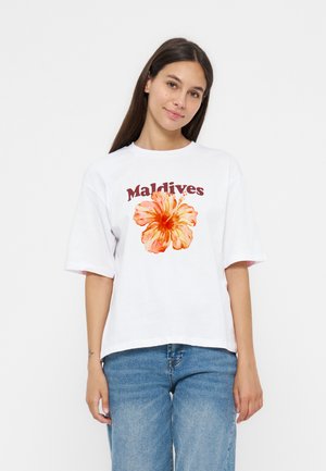 Jeune femme aux longs cheveux foncés portant un t-shirt blanc avec une grande fleur d’hibiscus orange et le mot Maldives, associé à un jean bleu.