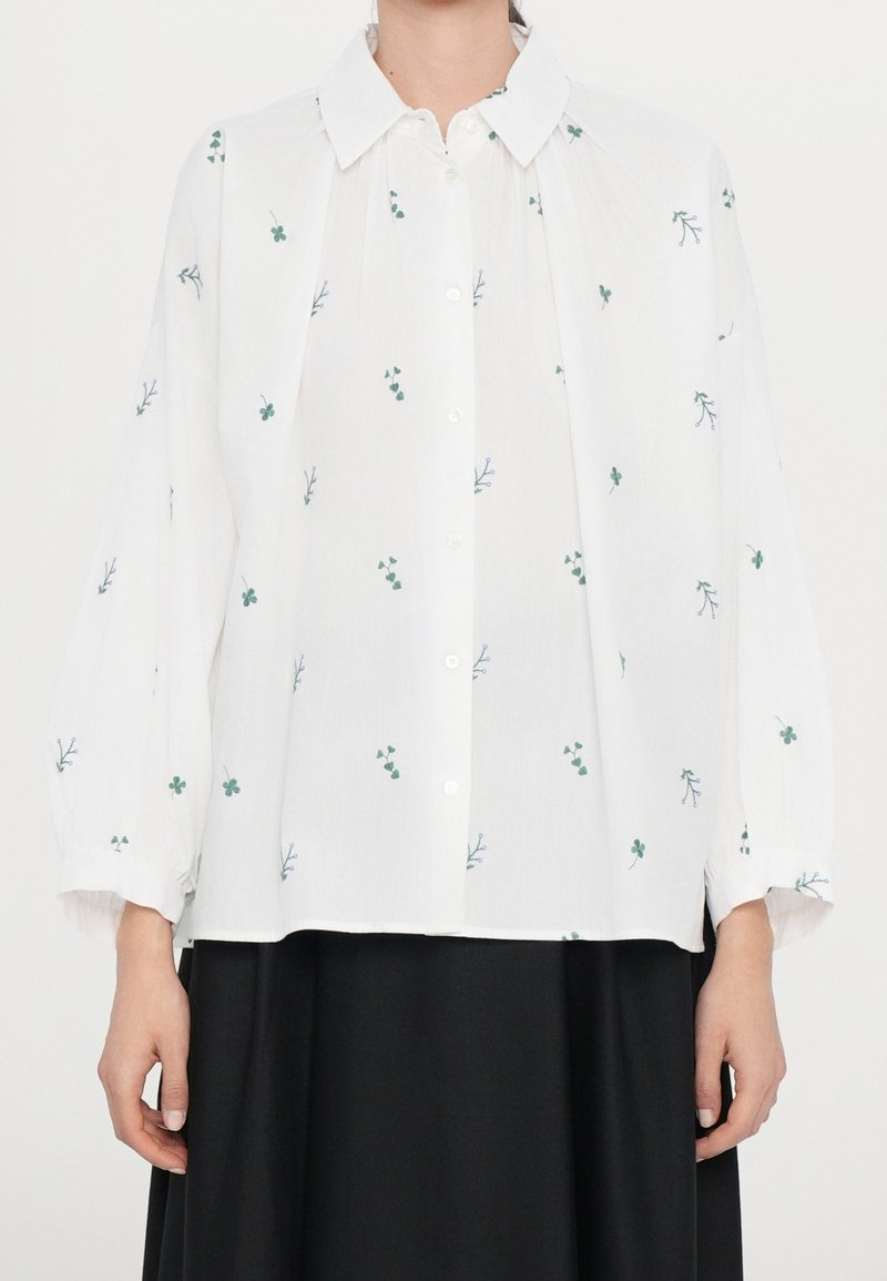 Blusa blanca de manga larga con bordado botánico verde, delante con botones, canesú plizado y mangas fruncidas. Llevada sobre una falda negra.