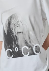 Balts T-krekls ar melnbaltu sievietes ar gariem matiem grafisko attēlu, virs kura uzdrukāts teksts "C°C" lieliem baltiem burtiem.